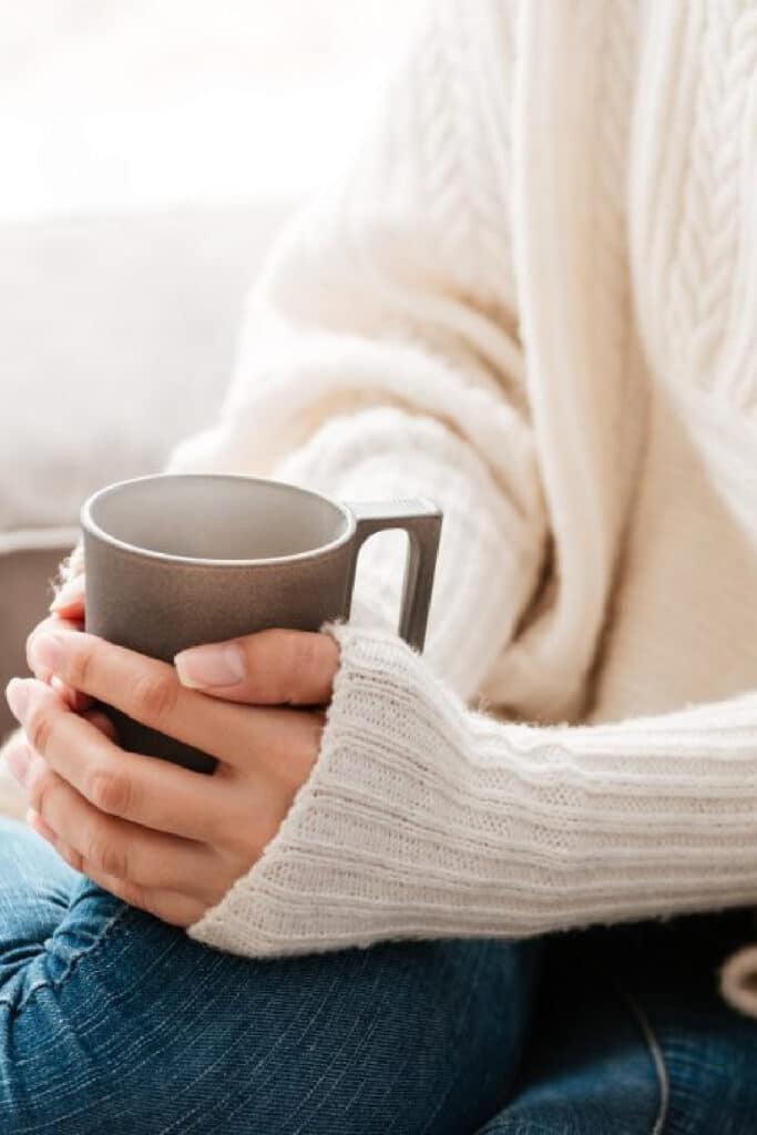 stay warm this winter - woman on couch warming hands of coffee mug